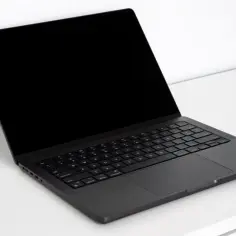 Ciemny laptop z czarnym ekranem, wyglądający jak Mac OS na PC, stoi na białym biurku.