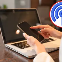 Dłonie trzymające smartfon nad klawiaturą laptopa, obok ikona Wi-Fi.