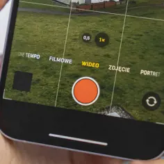Dłoń trzyma iPhone'a z otwartą aplikacją aparatu, widoczny przycisk nagrywania wideo.