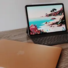 Złoty MacBook leży obok iPada z klawiaturą, który służy jako drugi ekran, wyświetlając kolorową tapetę z krajobrazem.