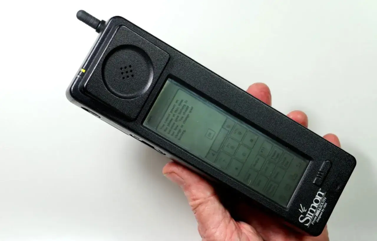 Dłoń trzyma czarny IBM Simon, pierwszy smartfon dotykowy, z komunikatem o niskim poziomie baterii na ekranie.