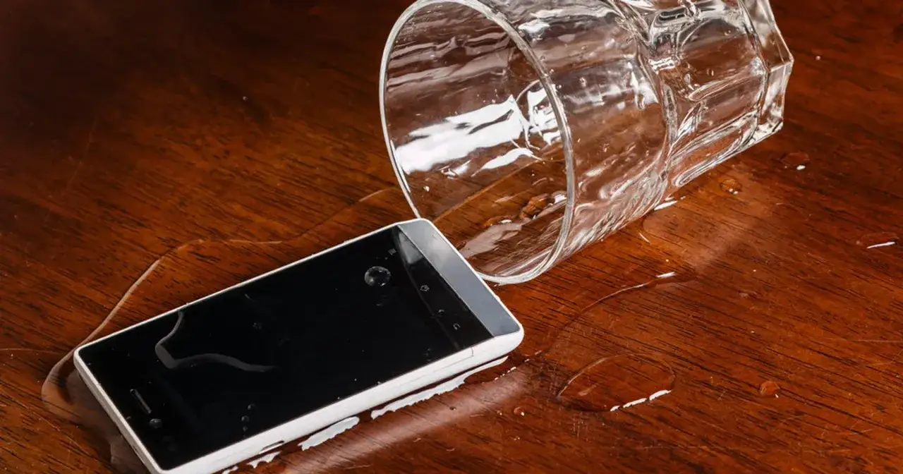 Szklanka z wodą przewrócona na drewniany stół, obok leży mokry iPhone. Wykryto ciecz iPhone.
